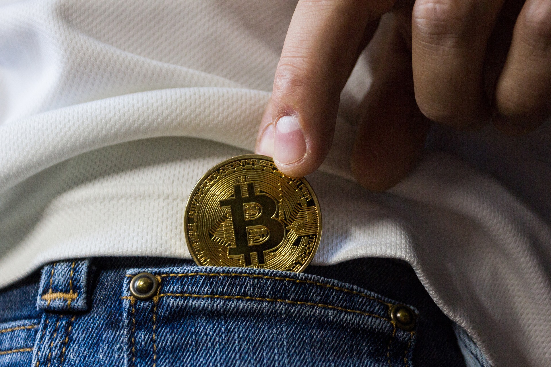 A hand slipping a gold Bitcoin coin into the pocket of a pair of jeans